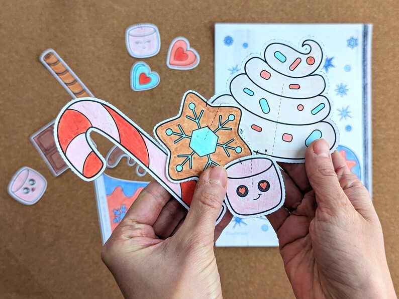 Peut inclure: Ensemble de d&eacute;coupes de papier sur le th&egrave;me de No&euml;l, comprenant une canne en bonbon, une &eacute;toile en pain d'&eacute;pices, une guimauve et une spirale de cr&egrave;me fouett&eacute;e avec des vermicelles. Les d&eacute;coupes sont tenues devant un livret partiellement visible.