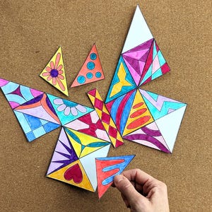 May include: A colorful butterfly puzzle made of triangular pieces. The puzzle pieces are decorated with various patterns and colors, including pink, blue, yellow, and red. A hand is holding one of the puzzle pieces.