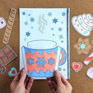 Peut inclure: Une page &agrave; colorier avec un motif de tasse, avec des flocons de neige et le texte "WINTER WARM UP". Autour de la tasse se trouvent des illustrations d&eacute;coup&eacute;es de guimauves, d'une barre chocolat&eacute;e, d'un biscuit, d'un sucre d'orge et d'autres &eacute;l&eacute;ments sur le th&egrave;me de l'hiver.