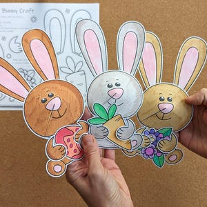 May include: Three colorful, cartoon bunny cutouts with pink-lined ears, holding a carrot, flowers, and an Easter egg. The bunnies are brown, gray, and tan. A sheet of paper with a bunny outline and the text "Spring Bunny Craft" is in the background.