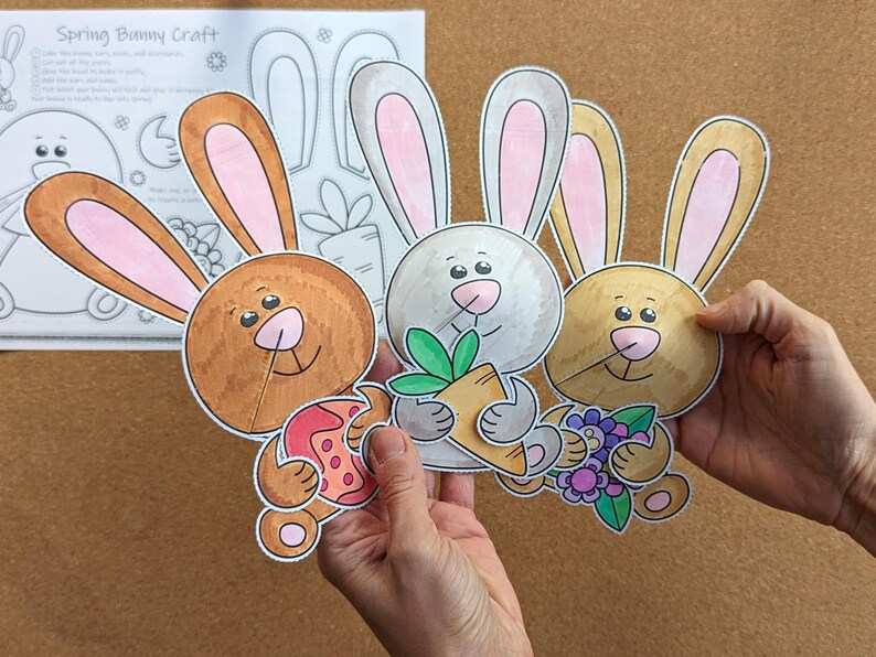 May include: Three colorful, cartoon bunny cutouts with pink-lined ears, holding a carrot, flowers, and an Easter egg. The bunnies are brown, gray, and tan. A sheet of paper with a bunny outline and the text "Spring Bunny Craft" is in the background.