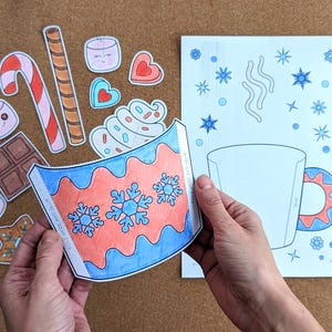 Peut inclure: Un projet de bricolage avec un motif de tasse en papier avec des flocons de neige bleus et rouges. L'image comprend des illustrations de nourriture en papier d&eacute;coup&eacute; comme des guimauves, des cannes en bonbon et du chocolat, sugg&eacute;rant un th&egrave;me hivernal.