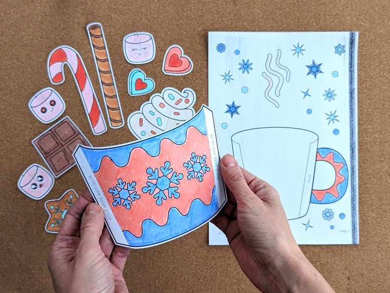 Peut inclure: Un projet de bricolage avec un motif de tasse en papier avec des flocons de neige bleus et rouges. L'image comprend des illustrations de nourriture en papier d&eacute;coup&eacute; comme des guimauves, des cannes en bonbon et du chocolat, sugg&eacute;rant un th&egrave;me hivernal.