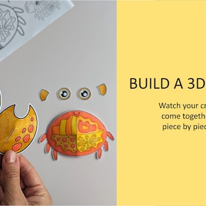 Op de afbeelding: Een papieren knutselset voor een 3D-krabproject met oranje en gele onderdelen met patronen, waaronder twee grote scharen, twee ogen en een lichaamsdeel. De tekst BUILD A 3D CRAB en Watch your crab come together piece by piece is zichtbaar.