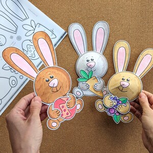 May include: Three colorful, cartoon bunny cutouts with pink-lined ears and smiling faces. One holds a carrot, another an Easter egg, and the third a bouquet of flowers. A sheet of bunny craft templates is in the background.