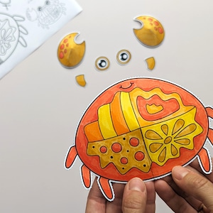 Op de afbeelding: Een papieren knutselset met een kleurrijke oranje en gele krab met gesegmenteerde patronen op het schild. De set bevat losse uitgesneden onderdelen voor de scharen en ogen van de krab, samen met een zwart-wit sjabloonblad.
