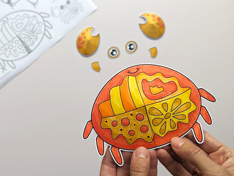 Op de afbeelding: Een papieren knutselset met een kleurrijke oranje en gele krab met gesegmenteerde patronen op het schild. De set bevat losse uitgesneden onderdelen voor de scharen en ogen van de krab, samen met een zwart-wit sjabloonblad.
