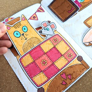 May include: A colorful, hand-drawn illustration of a cat, mouse, and bed. The cat is orange with blue eyes, and the mouse is gray. The bed has a pink and yellow quilt with flower details. The scene is part of a children's book or craft project.