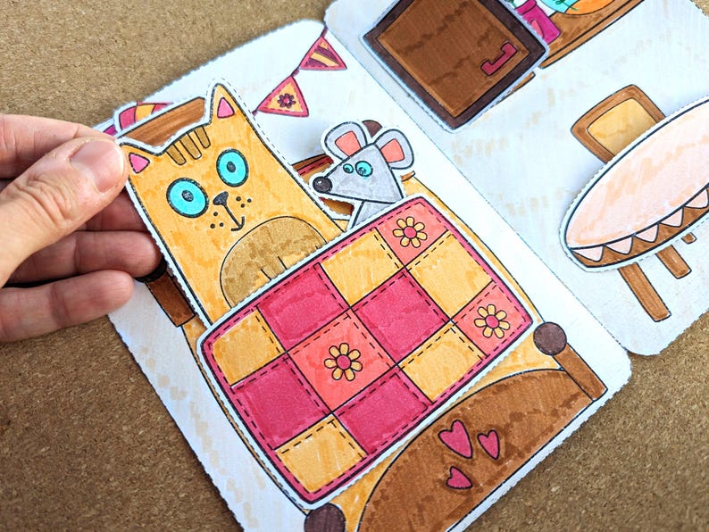 May include: A colorful, hand-drawn illustration of a cat, mouse, and bed. The cat is orange with blue eyes, and the mouse is gray. The bed has a pink and yellow quilt with flower details. The scene is part of a children's book or craft project.