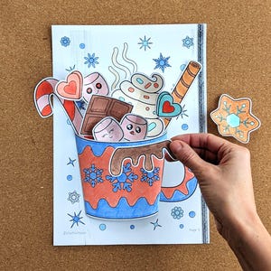 Peut inclure: Illustration color&eacute;e d'une tasse de chocolat chaud d&eacute;bordant de guimauves, de chocolat et d'un b&acirc;ton de sucre d'orge. La tasse est orn&eacute;e de flocons de neige et d'une anse. Un biscuit en pain d'&eacute;pices est &agrave; c&ocirc;t&eacute; de la tasse. L'image est sur fond blanc.