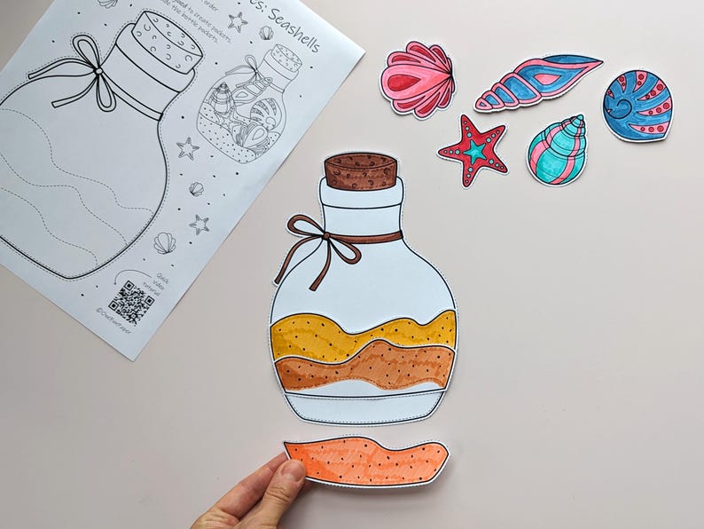 May include: A craft project featuring a jar illustration with a cork and a tied ribbon. The jar is filled with layers of yellow and brown. Several seashell cutouts in pink, blue, and green are scattered around the jar. A sheet of paper with the title "Seashells" is also present.