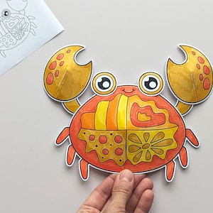 Op de afbeelding: Een papieren uitsnede van een krab met grote gele scharen en een rood lichaam verdeeld in vier secties, elk met verschillende patronen waaronder strepen, golvende lijnen, stippen en een bloem. De krab heeft twee grote ogen.