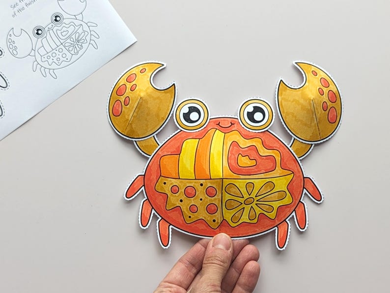Op de afbeelding: Een papieren uitsnede van een krab met grote gele scharen en een rood lichaam verdeeld in vier secties, elk met verschillende patronen waaronder strepen, golvende lijnen, stippen en een bloem. De krab heeft twee grote ogen.