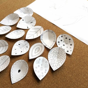 May include: A collection of 12 white paper leaves with black ink designs. Each leaf has a unique pattern, including swirls, dots, and lines.