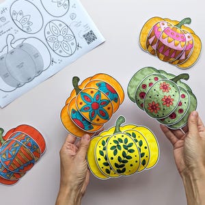 Op de afbeelding: Een papieren knutselproject met meerdere 3D-pompoenuitsparingen met kleurrijke, met de hand getekende patronen in oranje, geel, groen en roze. Een instructieblad getiteld Puffy Pumpkins Craft met diagrammen en een QR-code is zichtbaar.