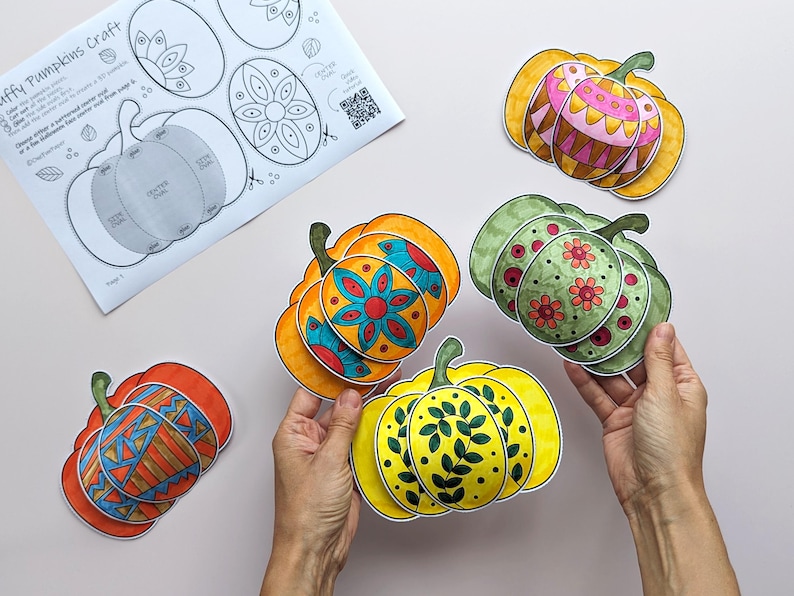 Op de afbeelding: Een papieren knutselproject met meerdere 3D-pompoenuitsparingen met kleurrijke, met de hand getekende patronen in oranje, geel, groen en roze. Een instructieblad getiteld Puffy Pumpkins Craft met diagrammen en een QR-code is zichtbaar.