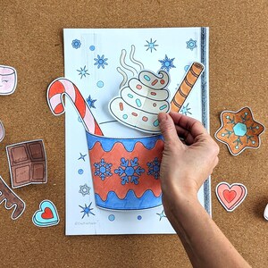Peut inclure: Une main positionne une d&eacute;coupe de cr&egrave;me fouett&eacute;e avec des vermicelles &agrave; c&ocirc;t&eacute; d'un dessin d'une tasse de chocolat chaud avec une canne en sucre et un b&acirc;ton de gaufrette. D'autres d&eacute;coupes incluent des guimauves, un biscuit en pain d'&eacute;pices et une barre de chocolat.