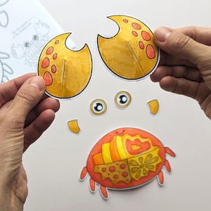 Op de afbeelding: Een papieren knutselset met een krab met een gesegmenteerd oranje en geel lichaam, twee gele scharen met oranje stippen en twee losse ogen. De onderdelen zijn voorzien van patronen en zijn bedoeld om uit te knippen en in elkaar te zetten.