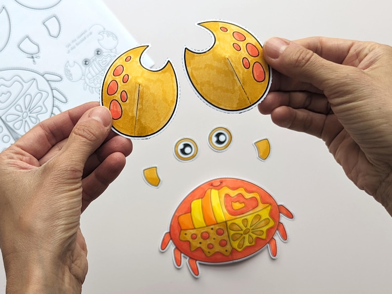 Op de afbeelding: Een papieren knutselset met een krab met een gesegmenteerd oranje en geel lichaam, twee gele scharen met oranje stippen en twee losse ogen. De onderdelen zijn voorzien van patronen en zijn bedoeld om uit te knippen en in elkaar te zetten.