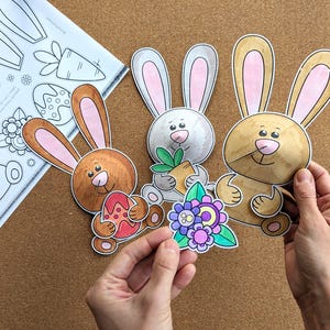 May include: Three colorful Easter bunny cutouts with pink-lined ears, holding an Easter egg, carrot, and flowers. A sheet of paper with bunny and carrot outlines is in the background. The cutouts are held by a person's hands.