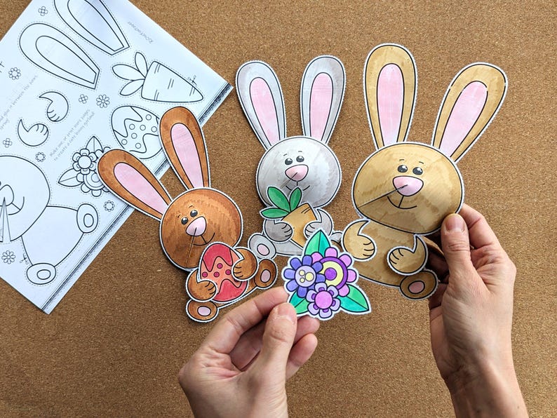 May include: Three colorful Easter bunny cutouts with pink-lined ears, holding an Easter egg, carrot, and flowers. A sheet of paper with bunny and carrot outlines is in the background. The cutouts are held by a person's hands.