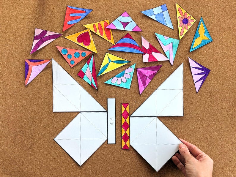 May include: A butterfly-shaped craft kit with colorful, geometric triangle pieces. The kit includes white paper shapes and smaller triangles with various designs, including hearts and flowers. The word "GLUE" is printed on one of the pieces.
