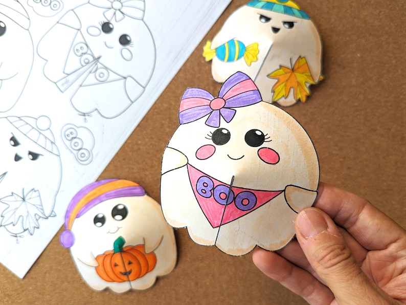 May include: Hand-colored paper ghost cutouts with cute faces and accessories. One ghost has a pink bandana with "BOO" on it. Other ghosts have hats, bows, and hold a pumpkin and candy. A sheet of ghost outlines is also visible.