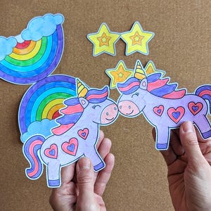 May include: Hand-drawn unicorn cutouts with heart details, rainbow, and star decorations. The unicorns have purple bodies, pink and purple manes, and yellow horns. The rainbow has multiple colours. The stars are yellow.