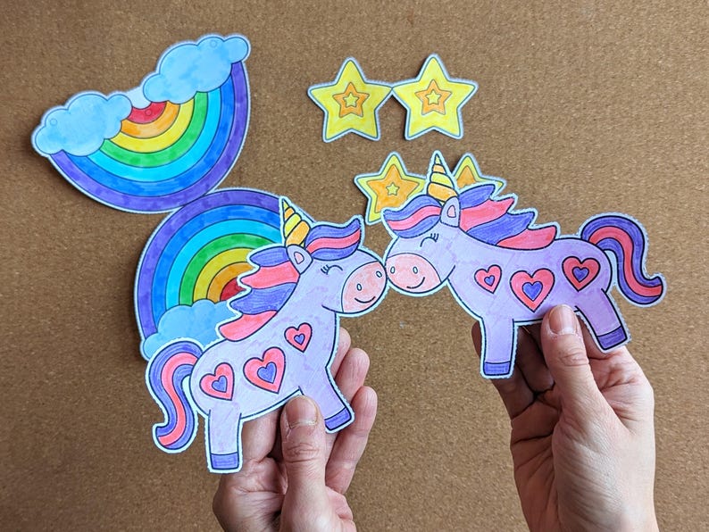 May include: Hand-drawn unicorn cutouts with heart details, rainbow, and star decorations. The unicorns have purple bodies, pink and purple manes, and yellow horns. The rainbow has multiple colours. The stars are yellow.