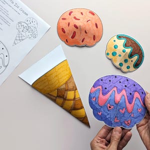 Op de afbeelding: Een kleurrijke, handgetekende ijshoorntjes knutselset. De set bevat een papieren hoorntje, drie bolletjes ijs in verschillende kleuren en een kleurplaat met instructies. Een bolletje wordt door een persoon vastgehouden.