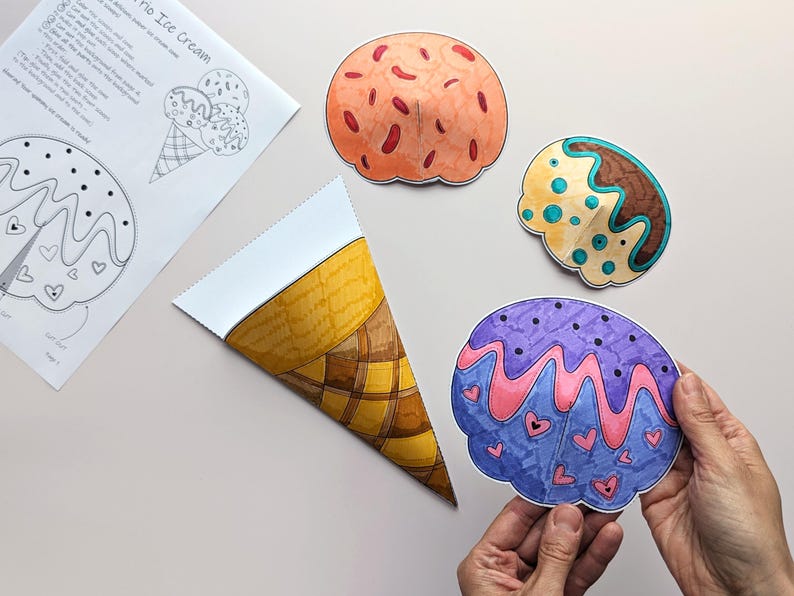 Op de afbeelding: Een kleurrijke, handgetekende ijshoorntjes knutselset. De set bevat een papieren hoorntje, drie bolletjes ijs in verschillende kleuren en een kleurplaat met instructies. Een bolletje wordt door een persoon vastgehouden.