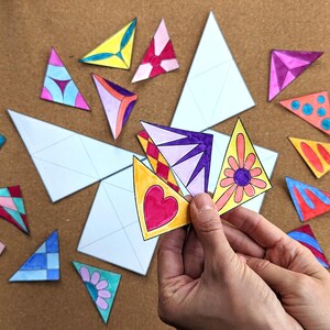 May include: A collection of colorful, hand-drawn triangular shapes with various designs, including hearts, flowers, and geometric patterns. Some triangles are blank, and others are held in a hand. The shapes are scattered on a brown surface.
