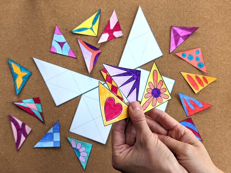 May include: A collection of colorful, hand-drawn triangular shapes with various designs, including hearts, flowers, and geometric patterns. Some triangles are blank, and others are held in a hand. The shapes are scattered on a brown surface.