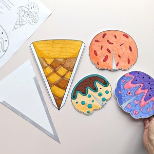 Op de afbeelding: Een verzameling kleurrijke, handgetekende ijsjeskegel- en bolvormige uitsparingen. De kegel is geel en bruin, terwijl de bollen oranje, bruin en blauw zijn met decoratieve patronen. Een blanco witte driehoek en een vel met ijsjeskegel-illustraties zijn ook aanwezig.