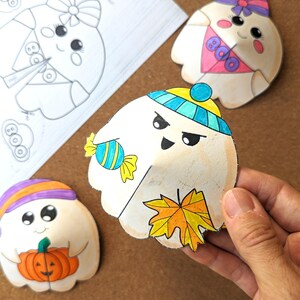 May include: Hand-colored paper cutouts of cartoon ghosts, each with unique accessories and expressions. One ghost holds a candy, another a pumpkin, and a third has a maple leaf. A sheet of ghost outlines is in the background.