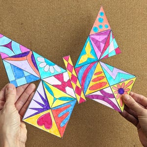 May include: A colorful, geometric butterfly design with various shapes and patterns. The wings feature triangles, squares, and other shapes filled with vibrant colors like pink, blue, yellow, and purple. The artwork is held by two hands.