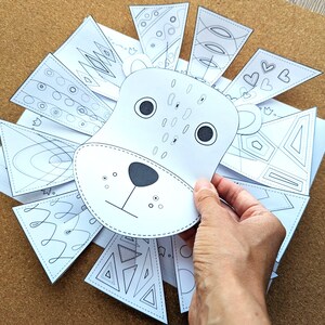 May include: A white paper lion craft with a hand holding the lion's face. The lion's mane is made of white paper triangles with various black line designs. The lion's face has black eyes, a nose, and whiskers.