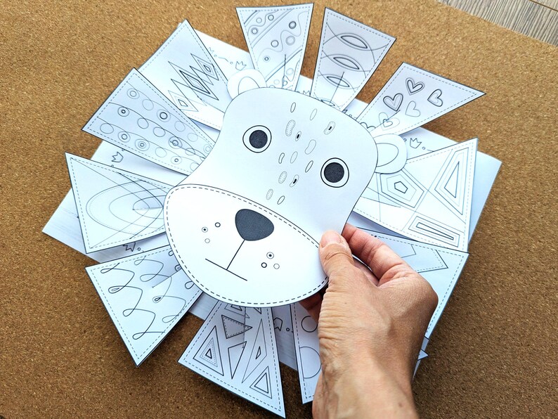 May include: A white paper lion craft with a hand holding the lion's face. The lion's mane is made of white paper triangles with various black line designs. The lion's face has black eyes, a nose, and whiskers.