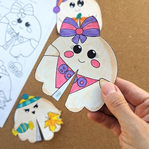 May include: A hand holds a cut-out ghost with a pink and purple bow, a pink bib with the word "BOO", and rosy cheeks. Other ghost cut-outs are visible, some colored and some uncolored, with different accessories. The theme is Halloween.