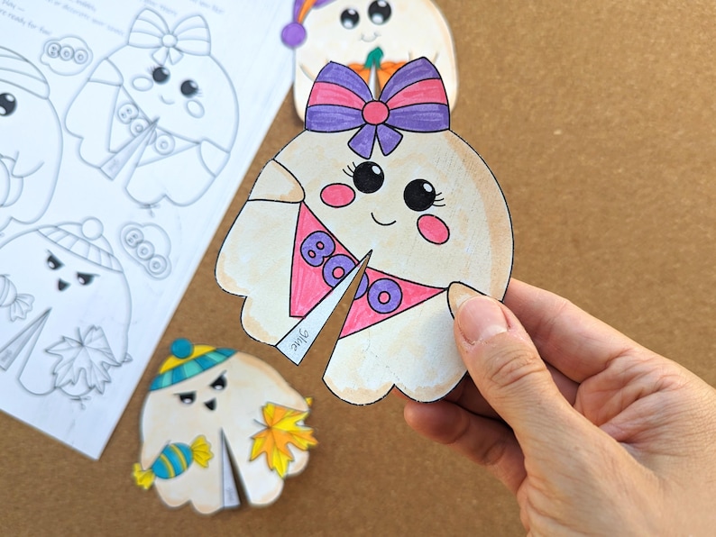 May include: A hand holds a cut-out ghost with a pink and purple bow, a pink bib with the word "BOO", and rosy cheeks. Other ghost cut-outs are visible, some colored and some uncolored, with different accessories. The theme is Halloween.