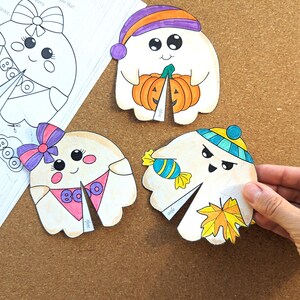 May include: Hand-drawn Halloween-themed ghost cutouts. One ghost holds a pumpkin, another has a candy, and a third has a fall leaf. The ghosts are white with colorful accents and the word "BOO" on one.