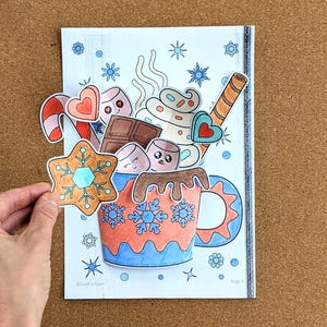 Peut inclure: Illustration color&eacute;e d'une tasse de chocolat chaud avec des guimauves, du chocolat et une canne en sucre. Une main tient un biscuit en pain d'&eacute;pices. La tasse est d&eacute;cor&eacute;e de flocons de neige. L'image est sur fond blanc.