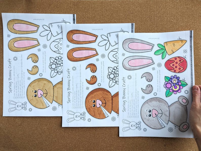 May include: Three sheets of paper with a spring bunny craft design. Each sheet features bunny ears, faces, and other spring-themed elements. The text "Spring Bunny Craft" is printed on each sheet. The sheets are on a brown surface.