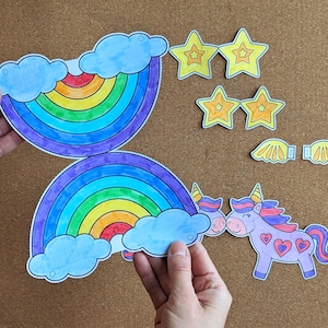 May include: Paper cutouts of a rainbow, stars, wings, and unicorns. The rainbow features blue clouds and a spectrum of colours. The unicorns are purple with pink manes and heart details. The stars and wings are yellow.