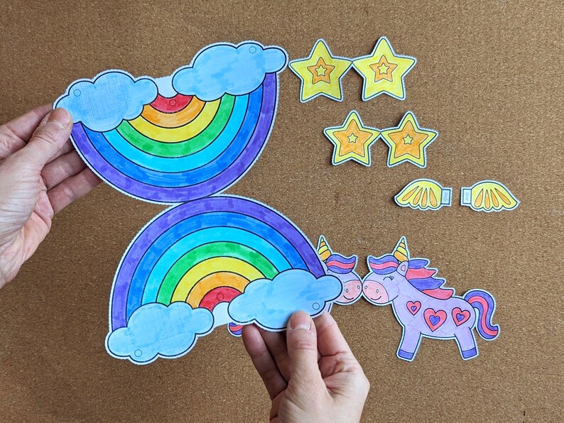 May include: Paper cutouts of a rainbow, stars, wings, and unicorns. The rainbow features blue clouds and a spectrum of colours. The unicorns are purple with pink manes and heart details. The stars and wings are yellow.
