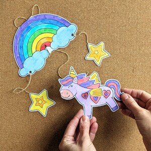 May include: A colourful, hand-drawn unicorn mobile featuring a rainbow, clouds, and stars. The unicorn is purple with a yellow horn and heart details. The rainbow has vibrant colours. The mobile is suspended by string.
