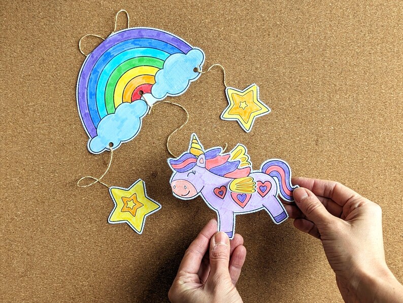 May include: A colourful, hand-drawn unicorn mobile featuring a rainbow, clouds, and stars. The unicorn is purple with a yellow horn and heart details. The rainbow has vibrant colours. The mobile is suspended by string.