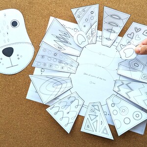 May include: A white paper craft kit featuring a bear face and various geometric shapes with black line art. The kit includes a bear face cutout and several patterned pieces to assemble. A hand is holding one of the pieces.