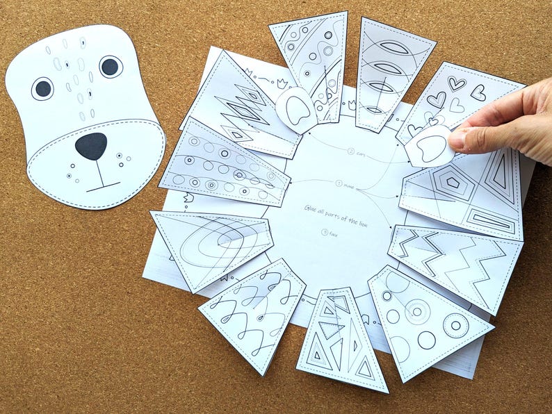 May include: A white paper craft kit featuring a bear face and various geometric shapes with black line art. The kit includes a bear face cutout and several patterned pieces to assemble. A hand is holding one of the pieces.