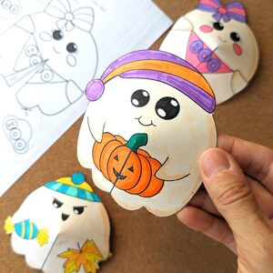 May include: Hand-drawn Halloween-themed cutouts of cartoon ghosts. One ghost holds an orange pumpkin, wearing a purple and orange hat. Other ghosts wear hats and bows. A sketch of similar ghosts is in the background.